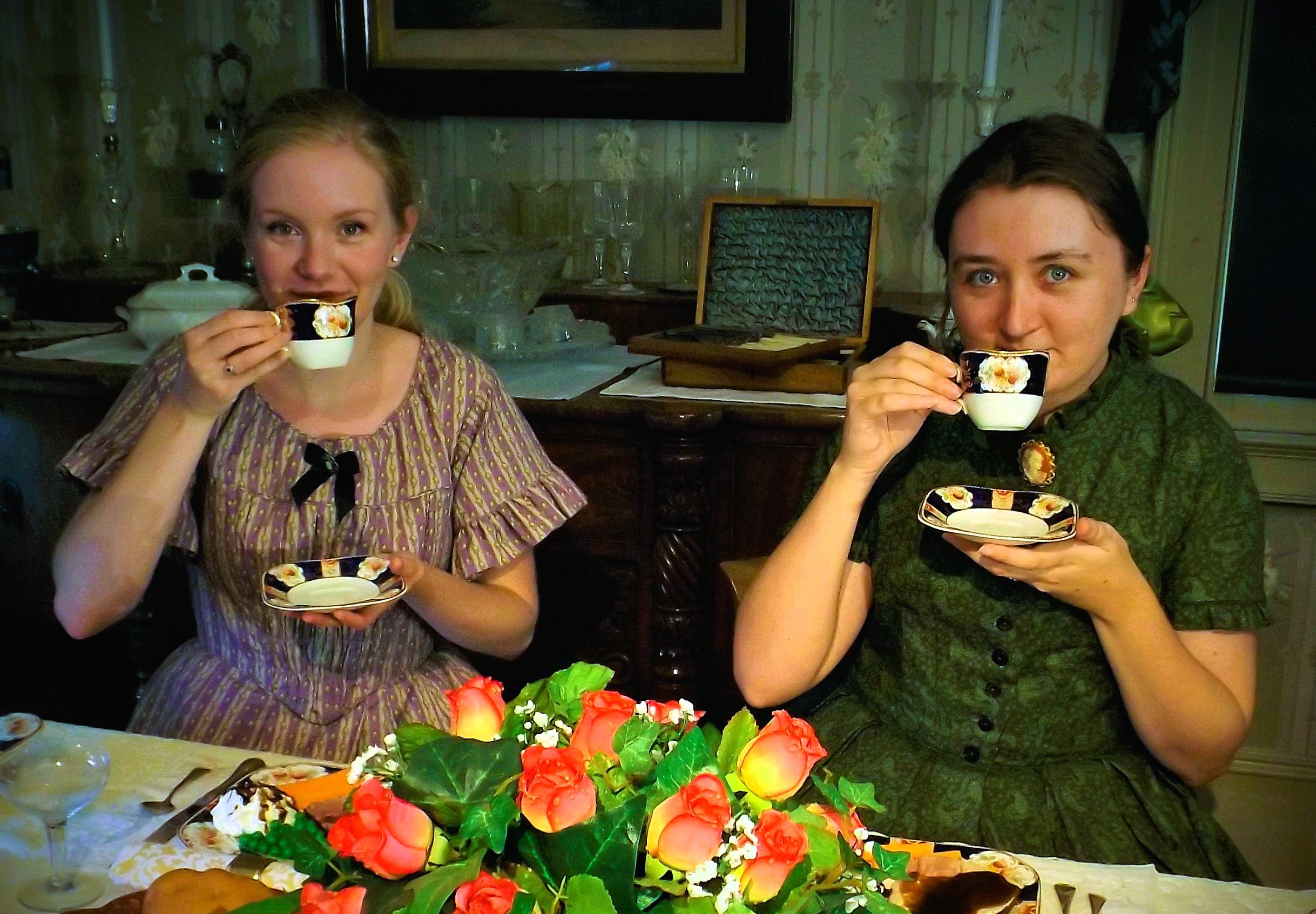 Two young ladies in Victorian dresses drink tea in the Lynde House dining room.